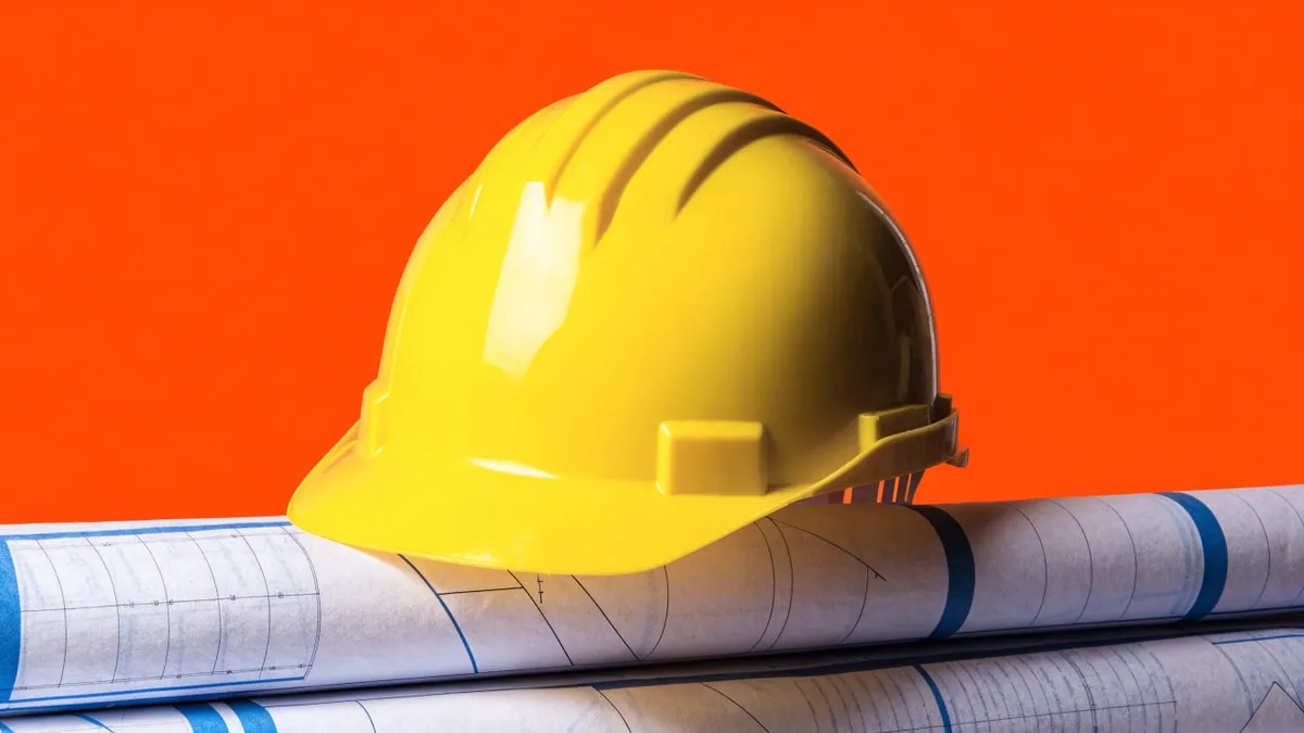 A yellow hard hat resting on a rolled set of site blueprints, cut out against a solid bright blue background.