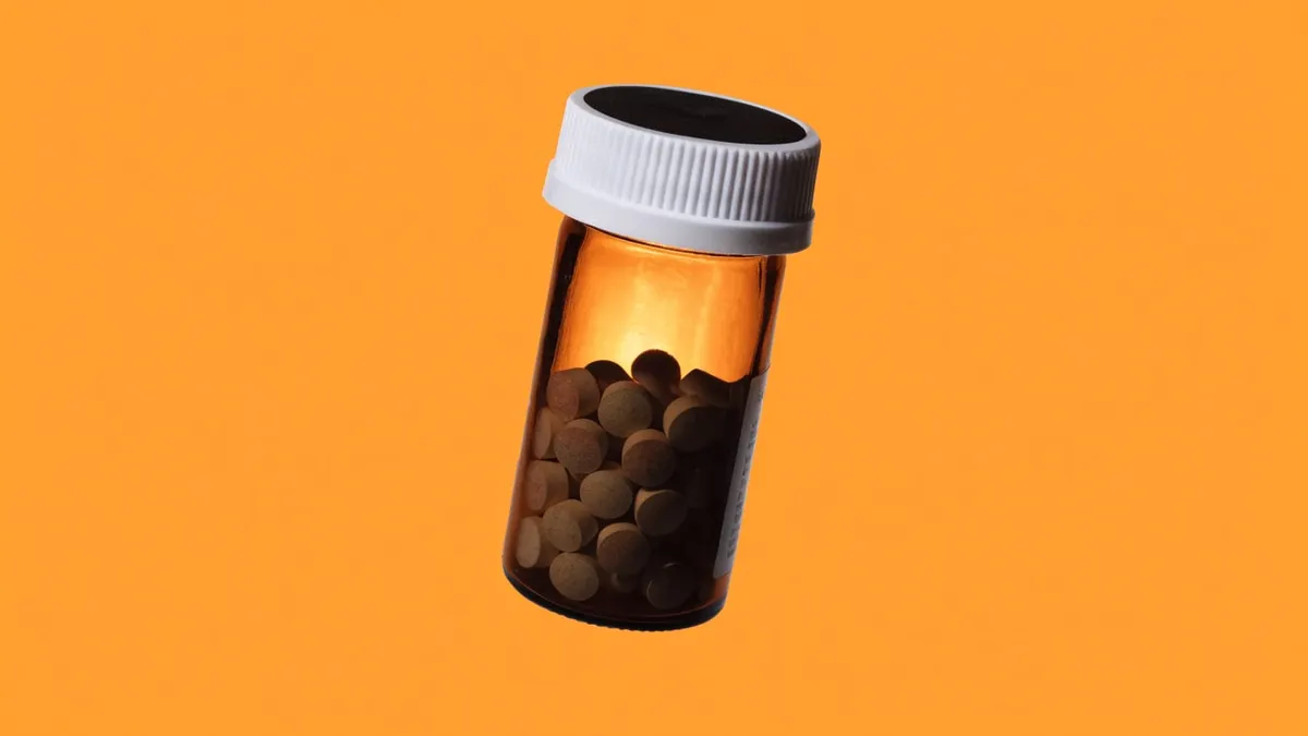A tilted amber prescription pill bottle with a white cap, filled with round tan tablets, cut out against a solid orange background.